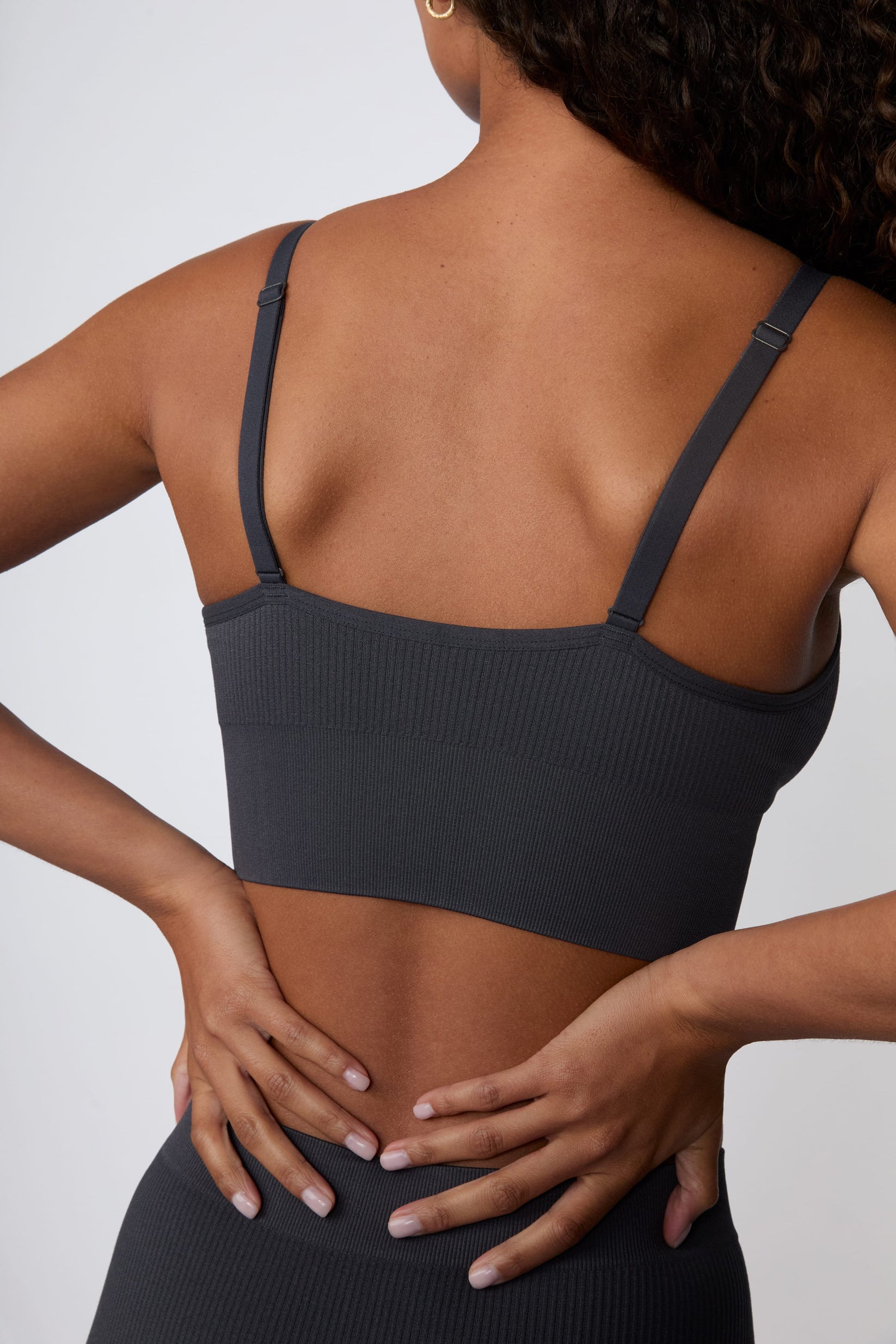 On a light gray studio backdrop, Courtney wears the Ribbed Seamless Triangle Bralette in Charcoal. A back view highlights the adjustable straps, ribbed texture, and wide underband.