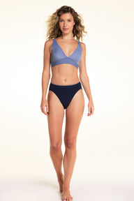 On a light studio backdrop, Alexis wears the Seamless Brief in Navy with a light blue bralette. The full-body image showcases the high-rise silhouette, smooth seamless construction, and contrasting waistband trim.