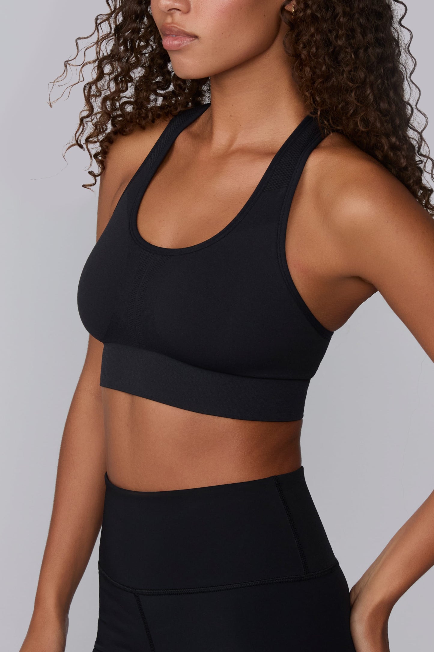 On a light gray studio backdrop, Courtney wears the Seamless Racerback Sports Bra in Black with matching bike shorts. The close-up view highlights the scoop neckline, wide underband, and seamless knit details through the front panel and straps.