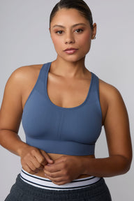 On a light gray studio backdrop, India wears the Seamless Racerback Sports Bra in Evernight with charcoal joggers. The close-up front view highlights the scoop neckline, textured knit detailing through the center panel, and supportive wide underband.