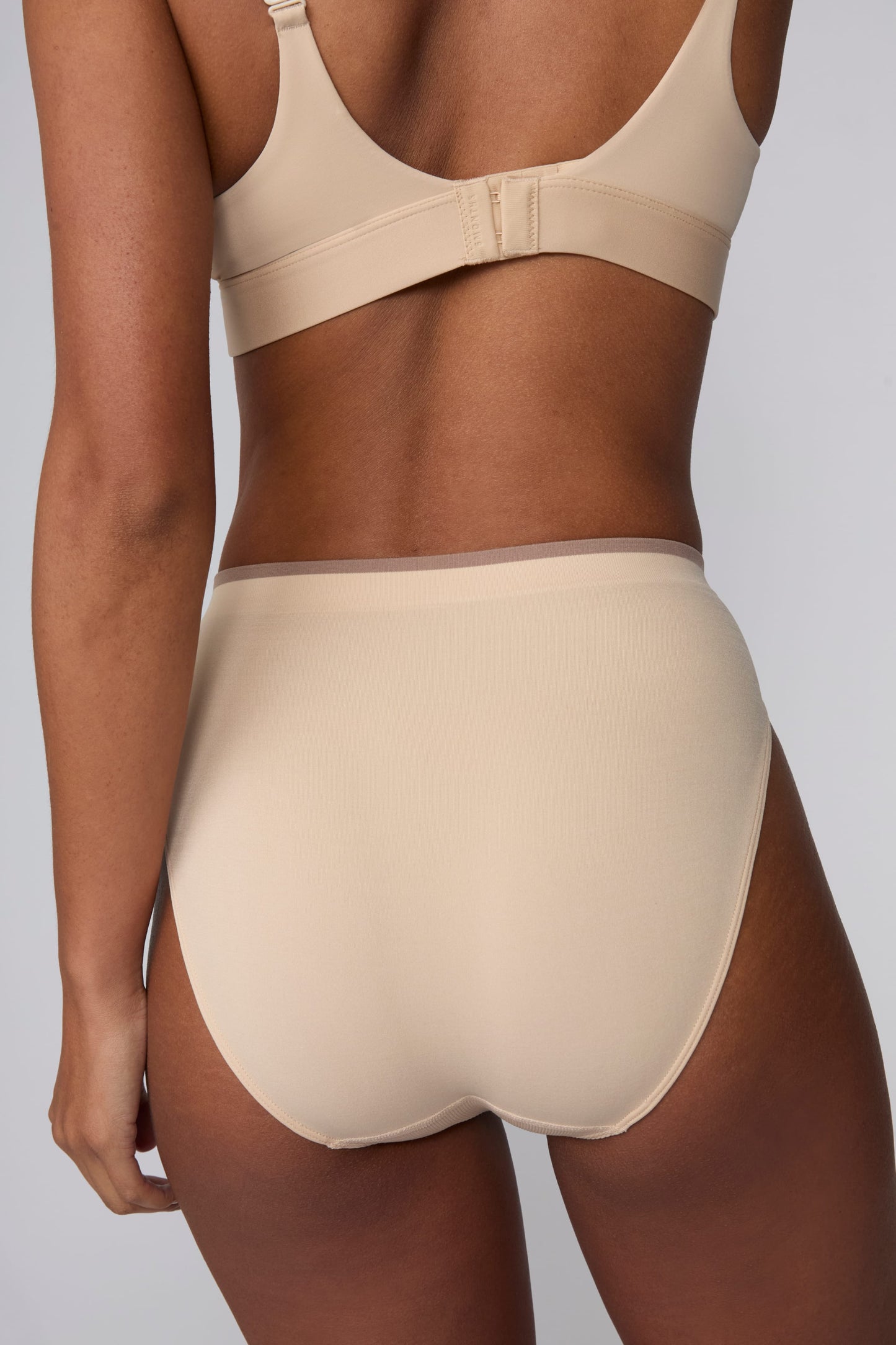 On a light gray studio backdrop, Courtney wears the Smooth Seamless Brief in Beige with the matching Smooth Seamless Bralette in Beige. The back view highlights the full back coverage, smooth finish, and contrast trim waistband.