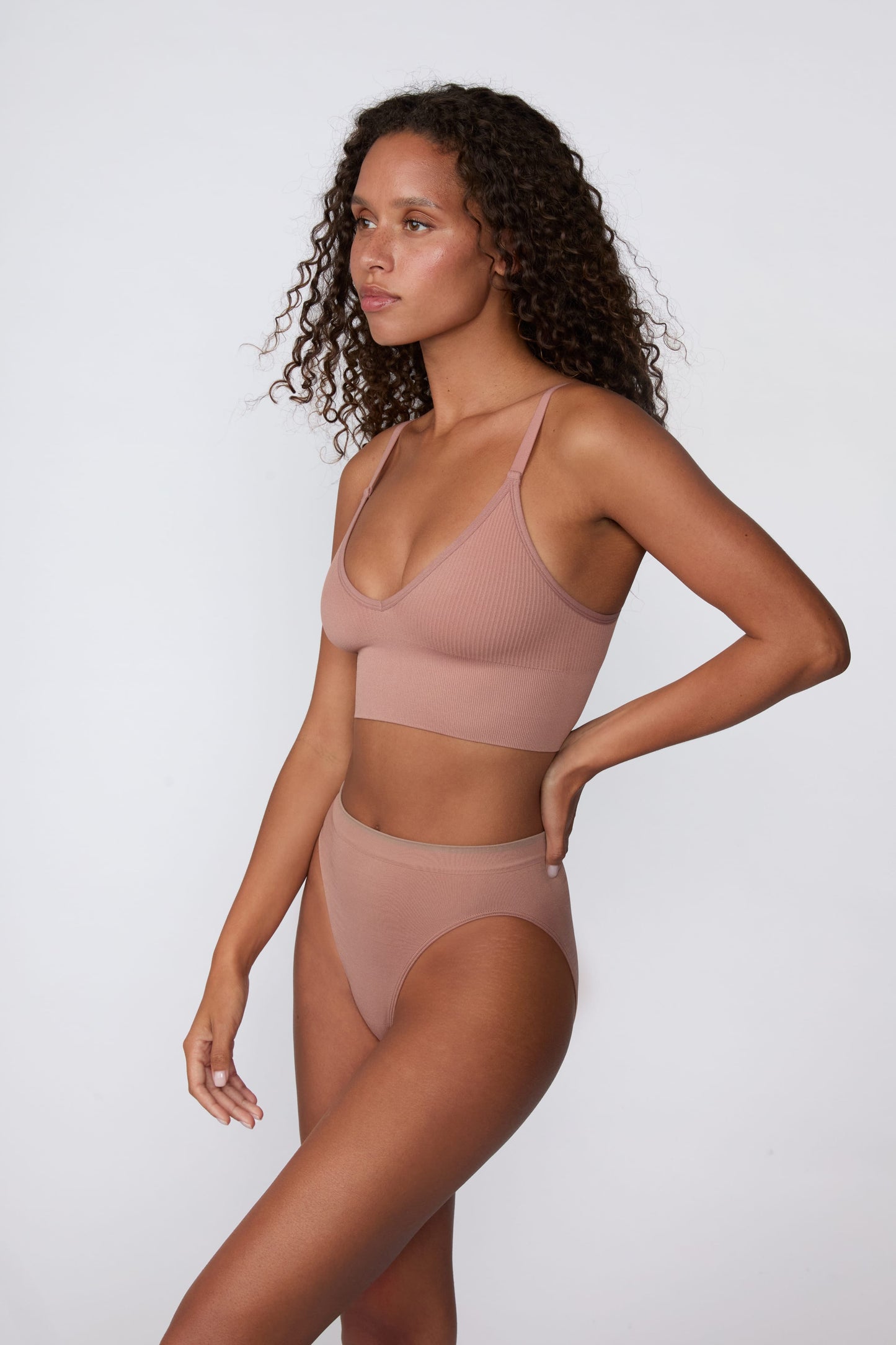 On a light gray studio backdrop, Courtney wears the Smooth Seamless Brief in Plum with the matching Seamless Scoop Bralette in Plum. The side view highlights the high-leg silhouette, smooth fabric, and contrast trim waistband.