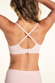 On a light neutral backdrop, Alexis is shown from the back wearing the Base Balconette Bra in Rose with the straps crossed. The image highlights the convertible crisscross straps, wide back band, and hook-and-eye closure.
