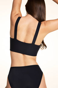 On a light neutral backdrop, Laura is shown from the back wearing the Bliss Smoothing Bralette in Black with matching underwear. The image highlights the straight back, wide straps, and smooth underband.
