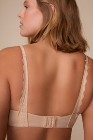 On a neutral studio backdrop, Sonnie wears the Base Lace T-Shirt Bra in Beige. The back view highlights the smooth band, adjustable straps, hook-and-eye closure, and lace racerback detail extending from the straps to the band.