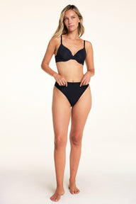 On a light studio backdrop, Meagan wears the Base Picot T-Shirt Bra in Black with matching black underwear. The full-body image highlights the smooth molded cups, picot trim along the neckline, and adjustable straps.