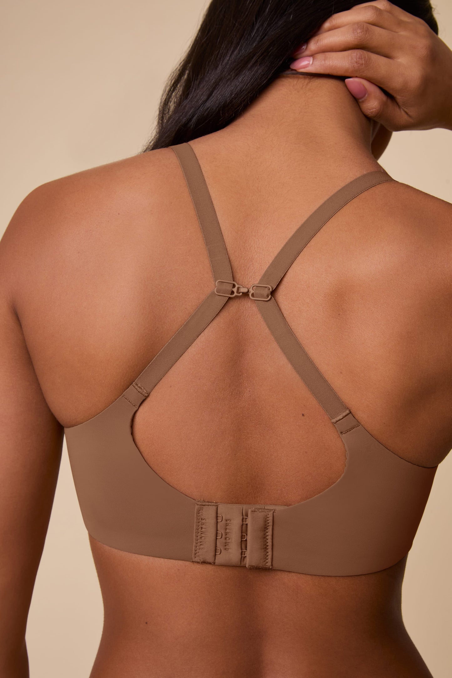 On a neutral beige backdrop, Ally wears the Base Unlined Bra in Brown with the J-hook fastened, creating a racerback silhouette and showcasing the convertible strap design.