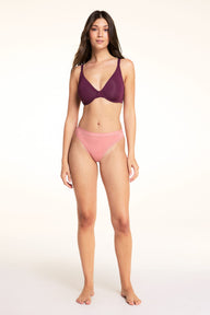 On a light studio backdrop, Laura wears the Base Unlined Bra in Eggplant. The full-body image showcases the unlined cups, underwire support, adjustable straps, and smooth silhouette.