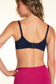 On a light studio backdrop, Alexis is shown from the back wearing the Base Unlined Bra in Navy with bright pink underwear. The image highlights the adjustable straps, hook-and-eye closure, and smooth back band.