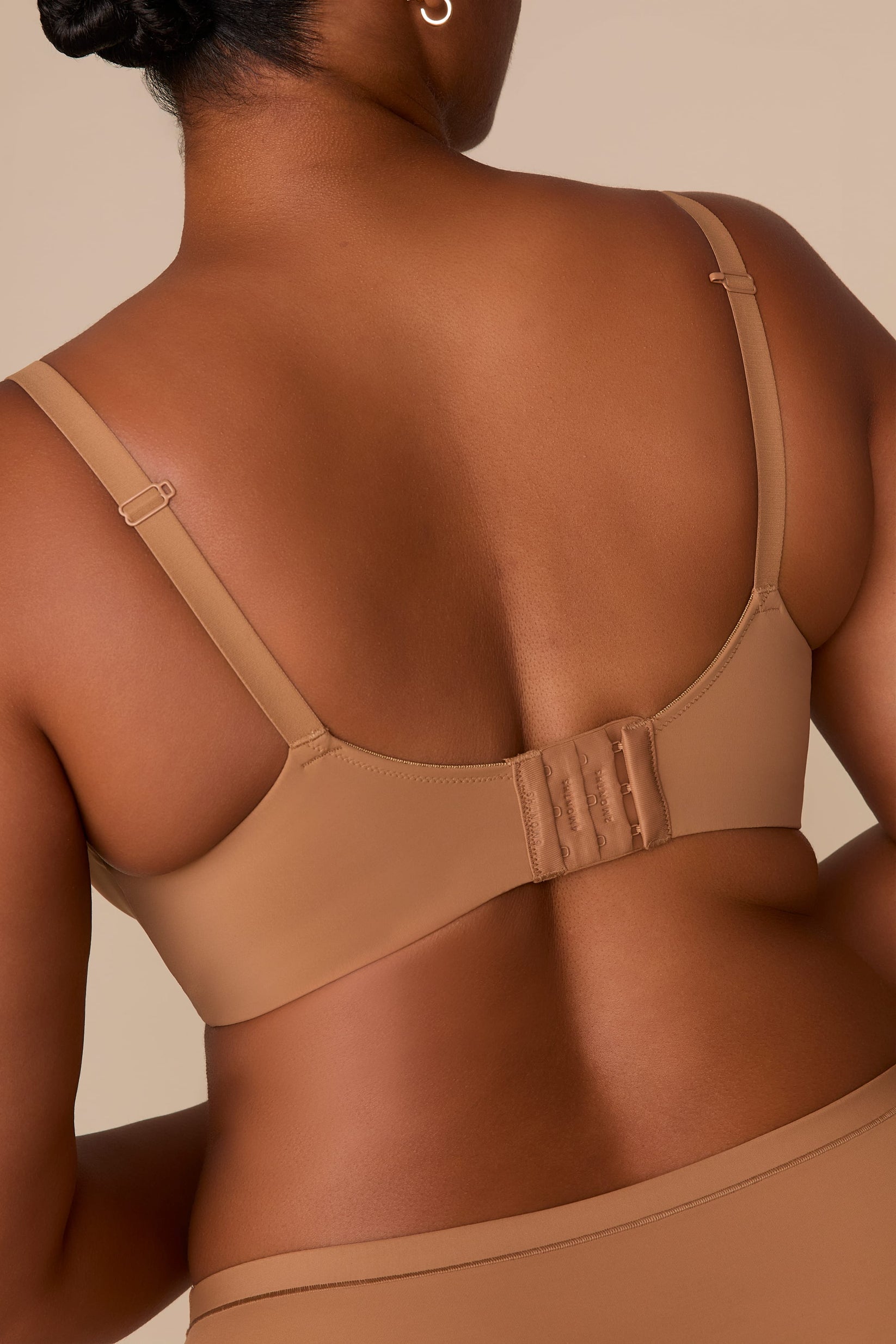 On a neutral beige backdrop, a back view of Zarria wearing the Base Wirefree T-Shirt Bra in Tan highlights the adjustable straps and hook-and-eye closure.