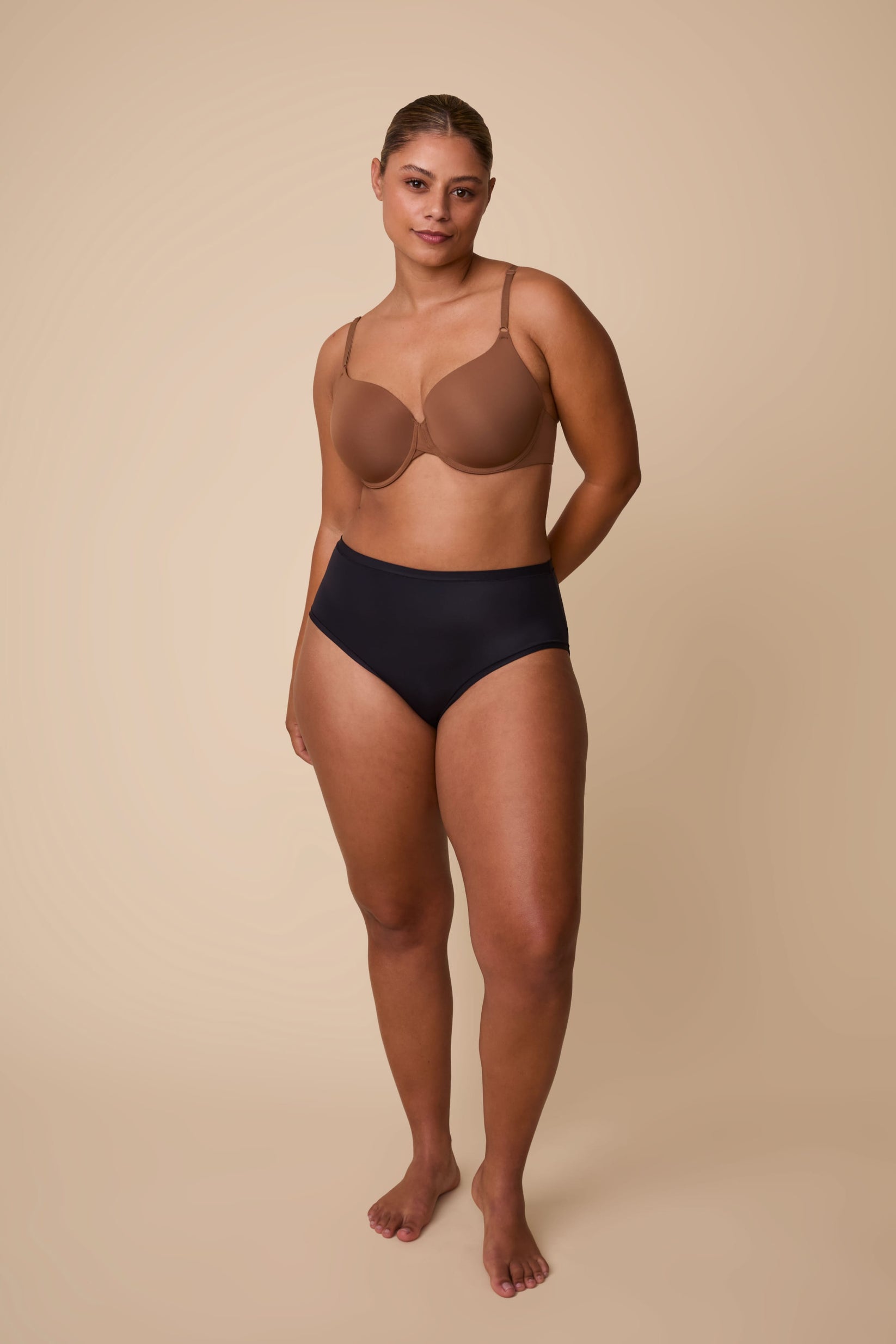On a neutral beige backdrop, India wears the Base T-Shirt Bra in Brown, standing in a full-body pose that showcases the bra’s smooth silhouette, lightly lined cups, and adjustable straps.