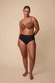 On a neutral beige backdrop, India wears the Base T-Shirt Bra in Brown, standing in a full-body pose that showcases the bra’s smooth silhouette, lightly lined cups, and adjustable straps.