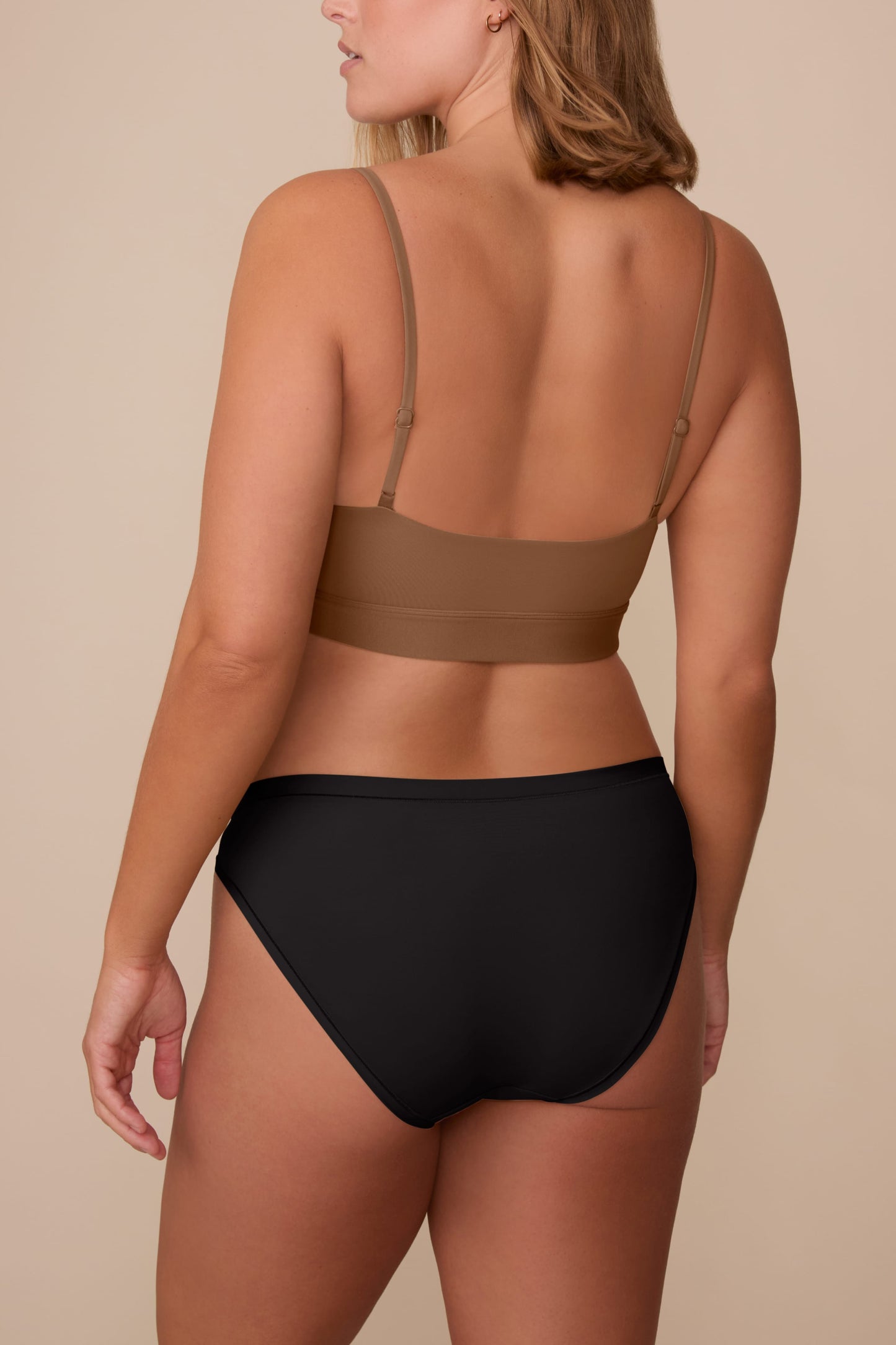 On a neutral beige backdrop, a back view of Sonnie wearing the Bliss Bikini in Black highlights the smooth finish and moderate back coverage.