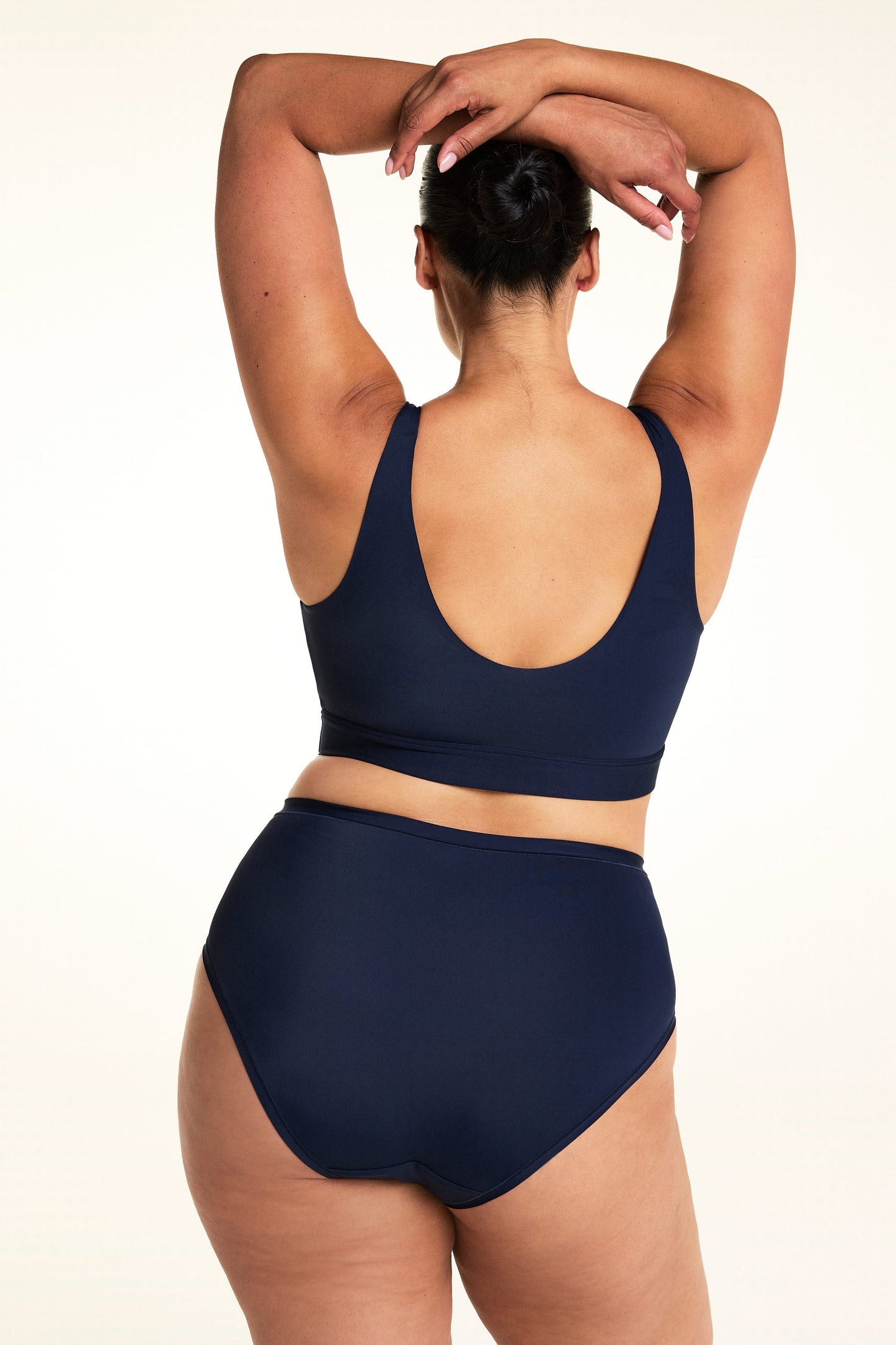On a light studio backdrop, Jennifer is shown from the back wearing the Bliss Highwaist in Navy with a matching navy bralette. With both arms raised, she showcases the full-coverage back and smooth, streamlined fit.