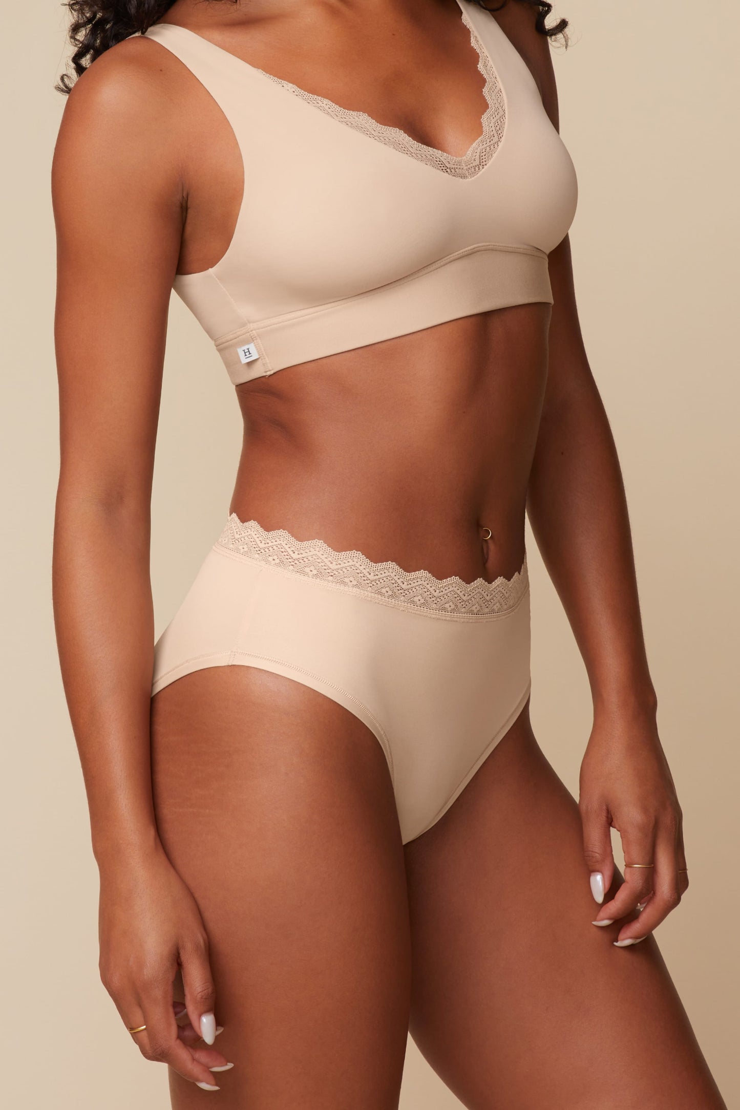 On a neutral beige backdrop, Ally wears the Bliss Lace Highwaist Brief in Beige with the matching Bliss Lace Bralette, highlighting the high-rise silhouette, smooth fabric, and lace waistband from a side angle.