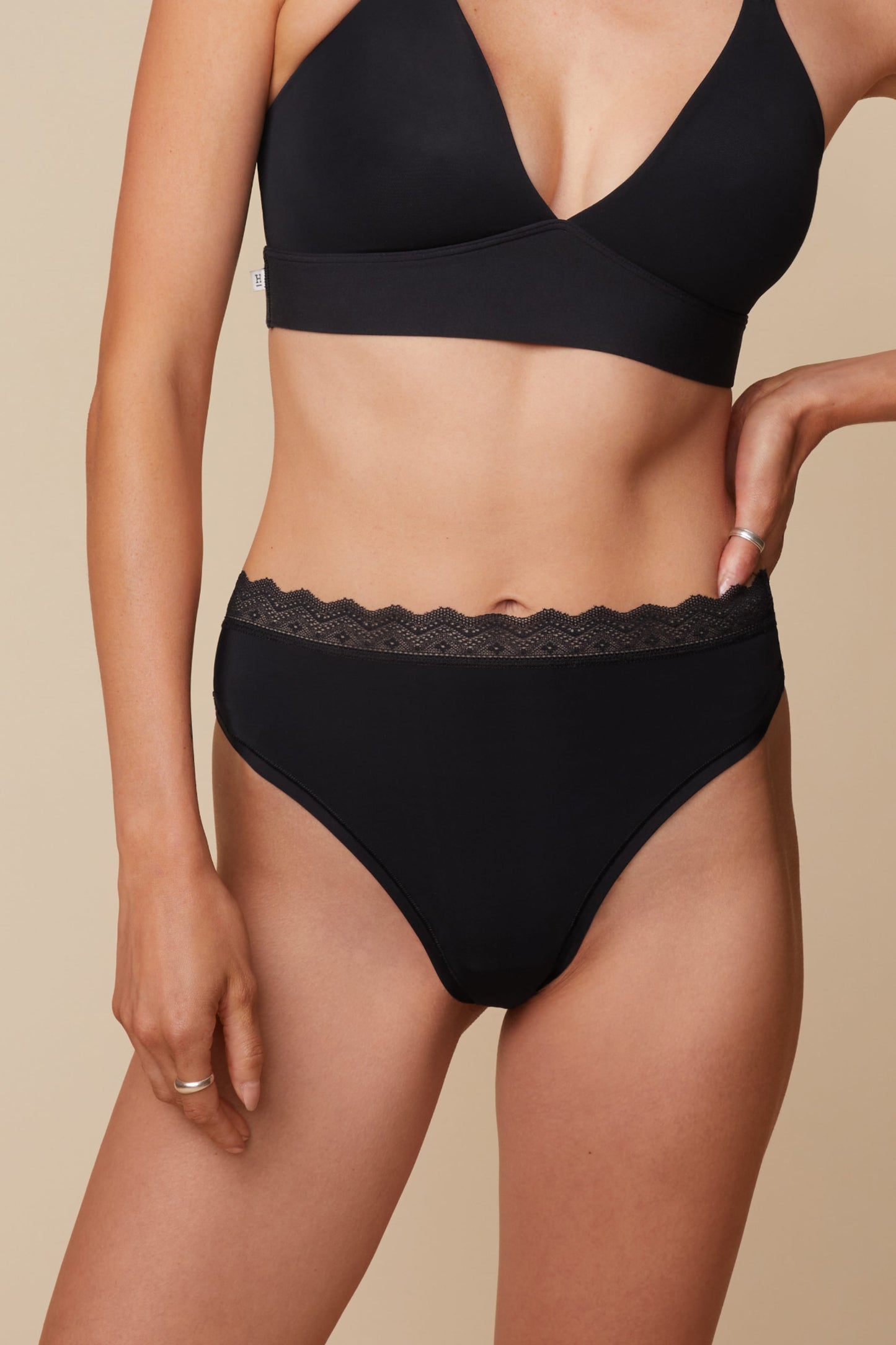 On a neutral beige backdrop, Anna wears the Bliss Lace Highwaist Thong in Black with the matching Bliss Lace Bralette, highlighting the smooth fabric, high-rise silhouette, and delicate lace waistband from a front angle.