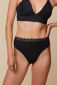 On a neutral beige backdrop, Anna wears the Bliss Lace Highwaist Thong in Black with the matching Bliss Lace Bralette, highlighting the smooth fabric, high-rise silhouette, and delicate lace waistband from a front angle.