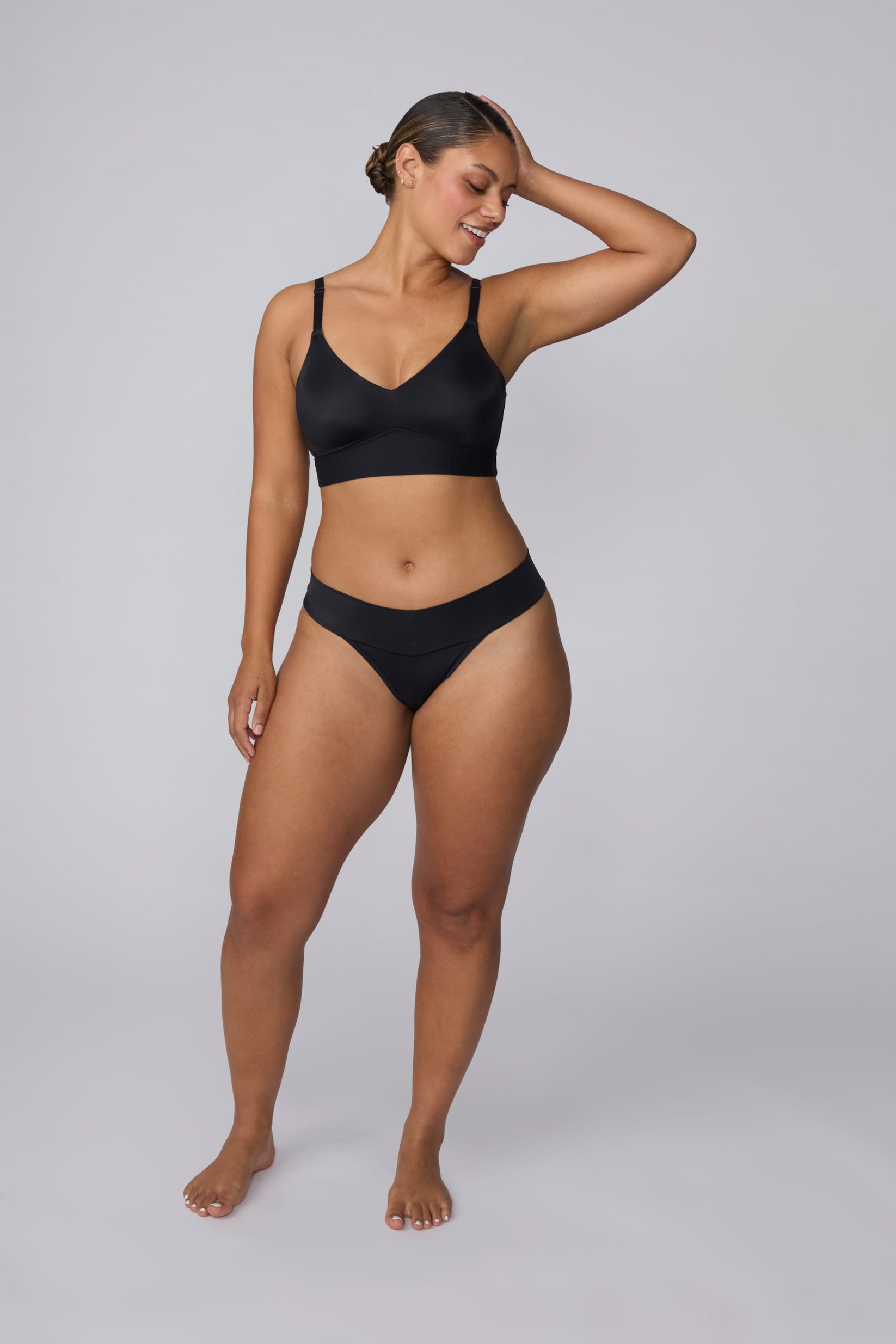 In a studio setting, India wears the Bliss Low Rise Thong in Black with the matching Bliss Bralette in Black. The full-body front view highlights the low-rise waistband, smooth fabric, and minimal-coverage thong silhouette.