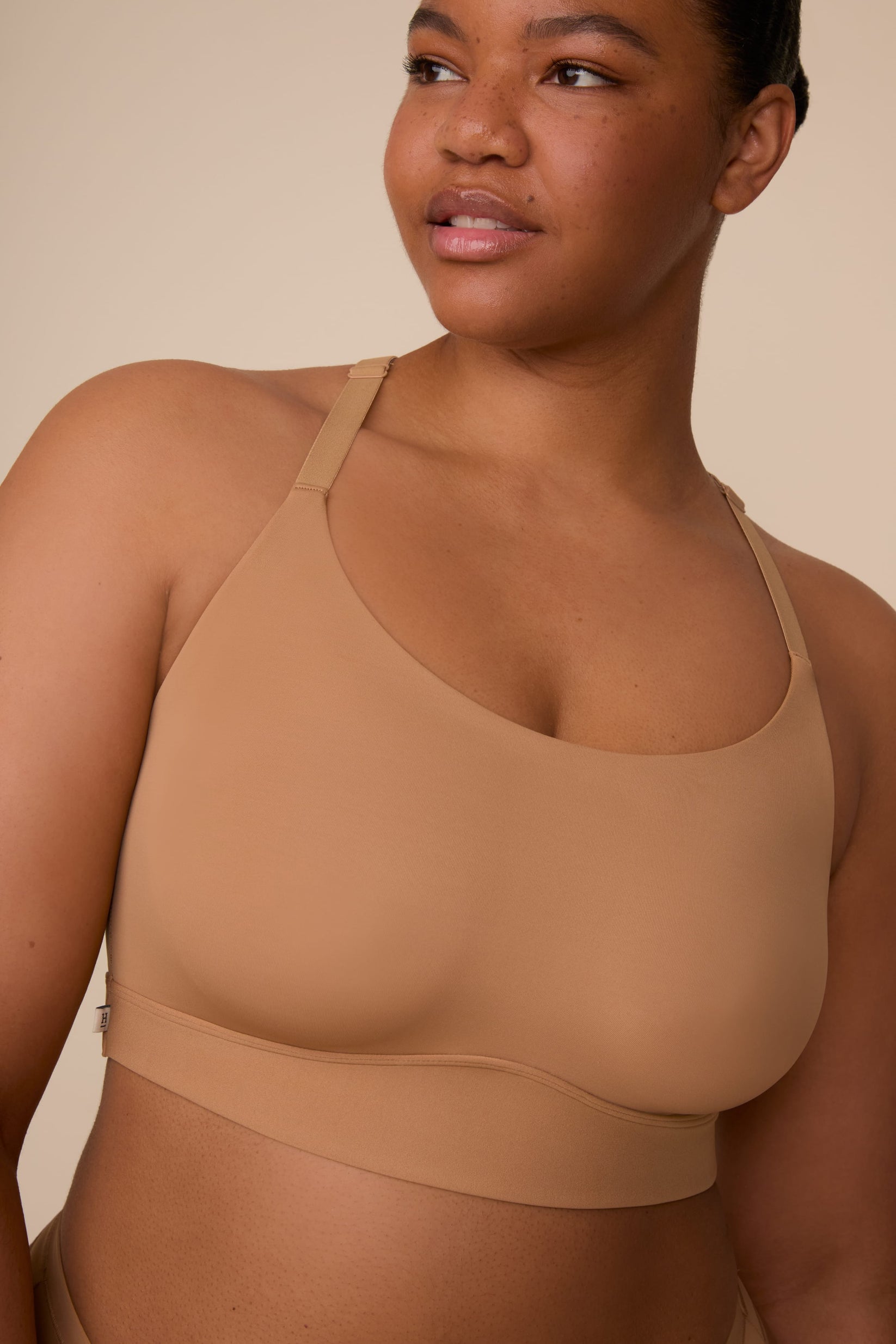 On a neutral beige backdrop, a close-up of Zarria wearing the Bliss Racerback Bralette in Tan showcases the smooth fabric, scoop neckline, and adjustable straps.