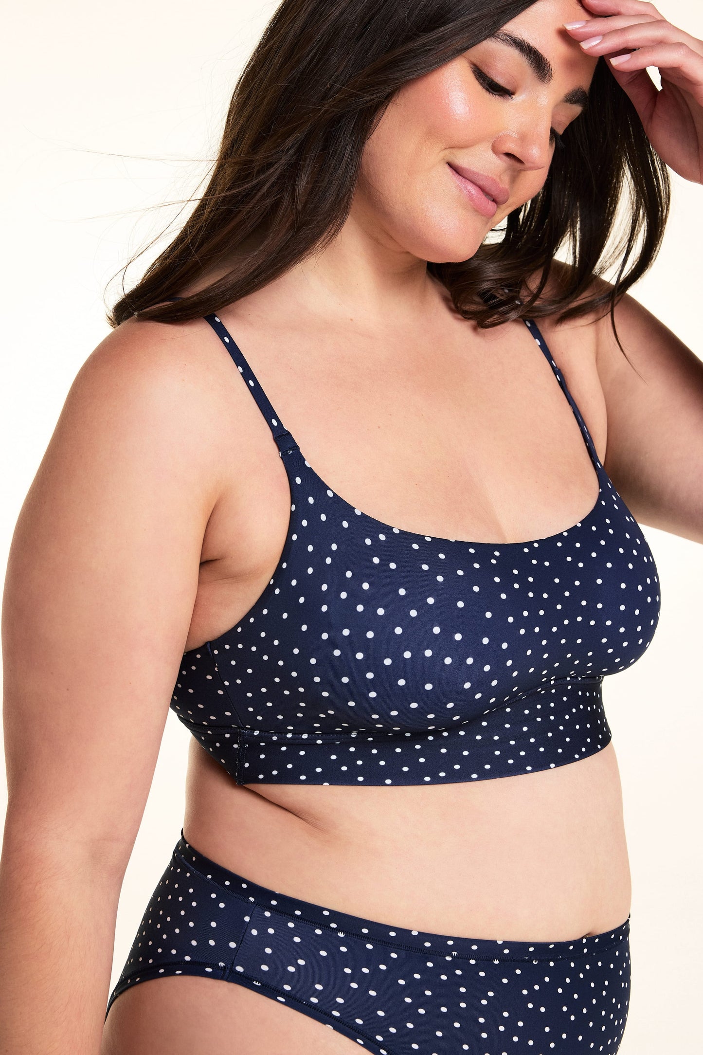 On a light studio backdrop, Alessandra wears the Bliss Scoop Bralette in Navy Dot with matching Navy Dot underwear. The angled close-up highlights the scoop neckline, adjustable straps, wide underband, and allover white dot print.