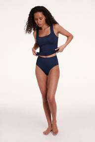 On a light studio backdrop, Courtney wears the Bliss Tank in Navy with matching Navy underwear. The full-body image shows her gently holding the hem, highlighting the square neckline and streamlined silhouette.