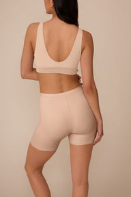 On a neutral studio backdrop, Marcela wears the Bliss Under Short in Beige with a matching beige bralette. The back view showcases the smooth waistband, full back coverage, and thigh-length design.