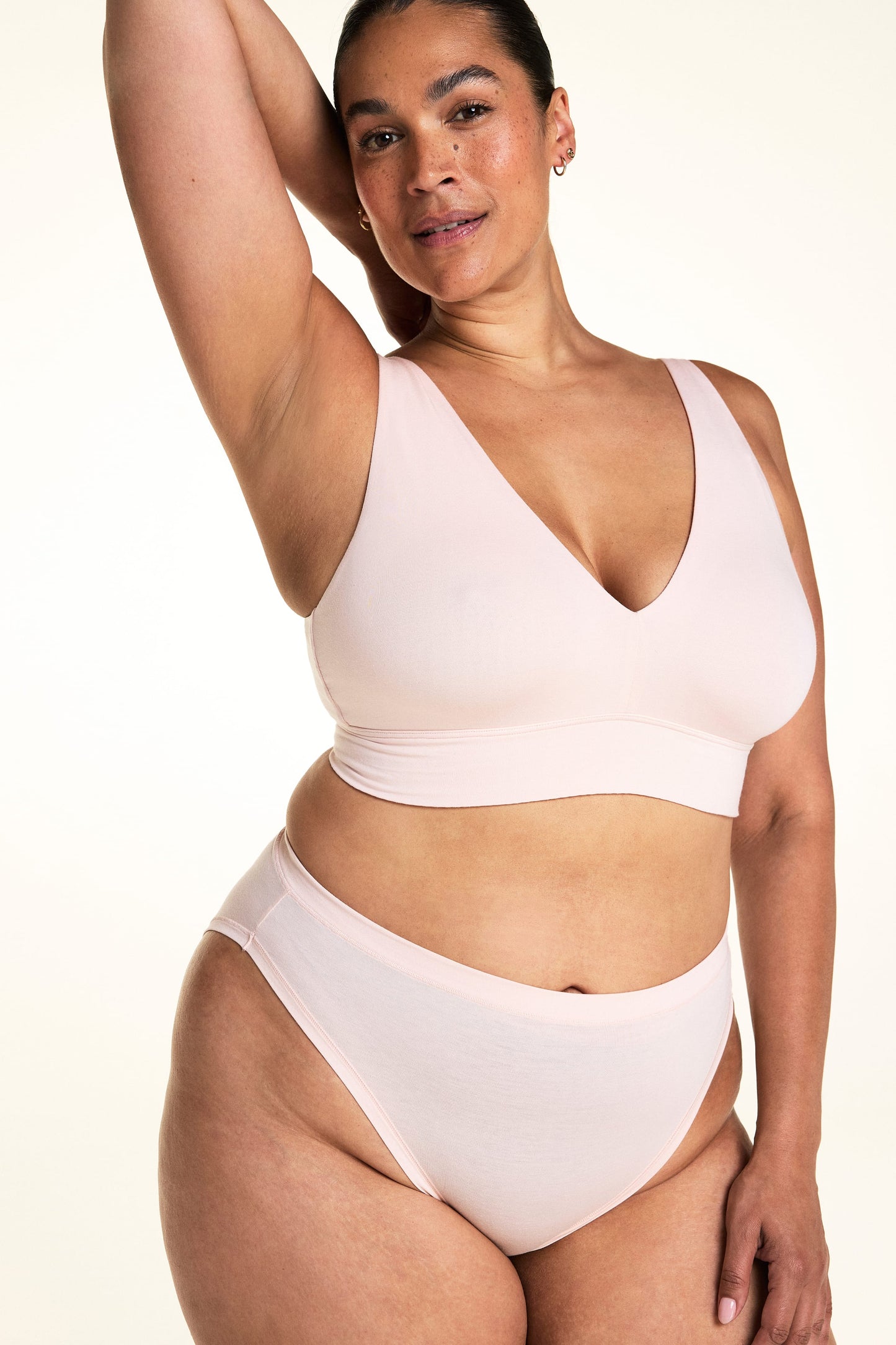 On a light studio backdrop, Jennifer wears the Cloud Cotton French Cut in Dogwood with the Bliss Triangle Bralette in Dogwood. The close-up highlights the deep V-neckline of the bralette, the high-cut leg opening, and the soft cotton fabric.