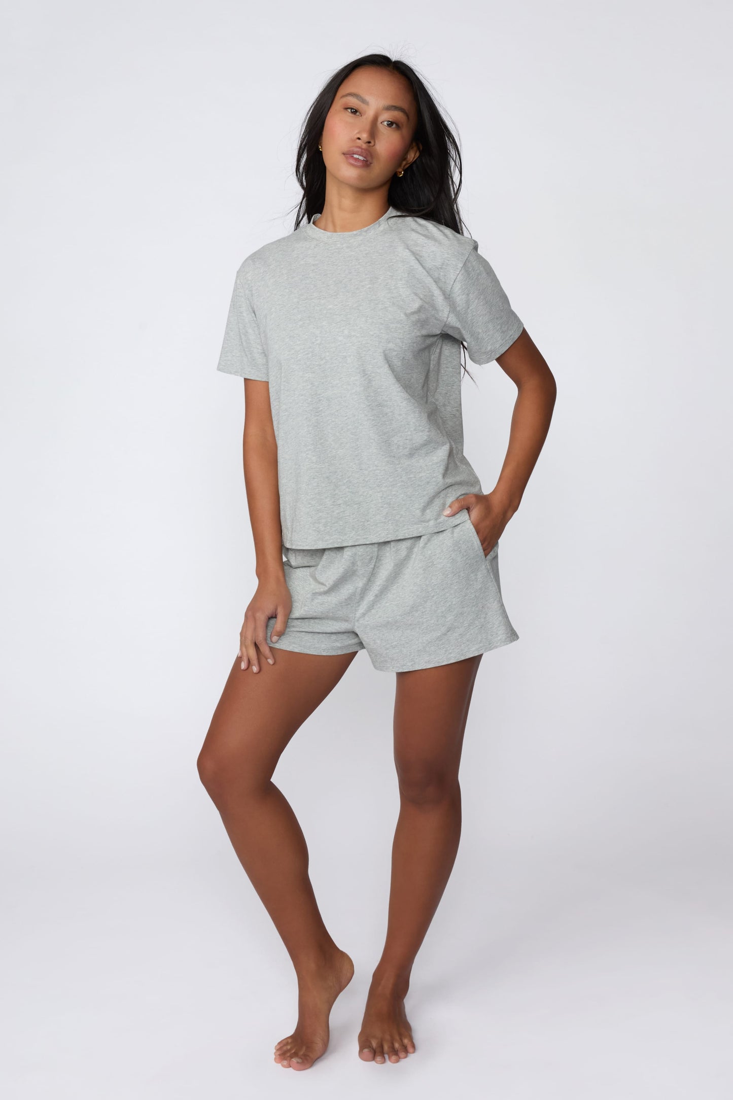 On a light gray studio backdrop, Amber wears the Cloud Cotton Pajama Shirt in Heather Grey with matching pajama shorts. The full-body view showcases the relaxed fit, crew neckline, and easy sleepwear styling.