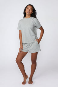 On a light gray studio backdrop, Amber wears the Cloud Cotton Pajama Shirt in Heather Grey with matching pajama shorts. The full-body view showcases the relaxed fit, crew neckline, and easy sleepwear styling.