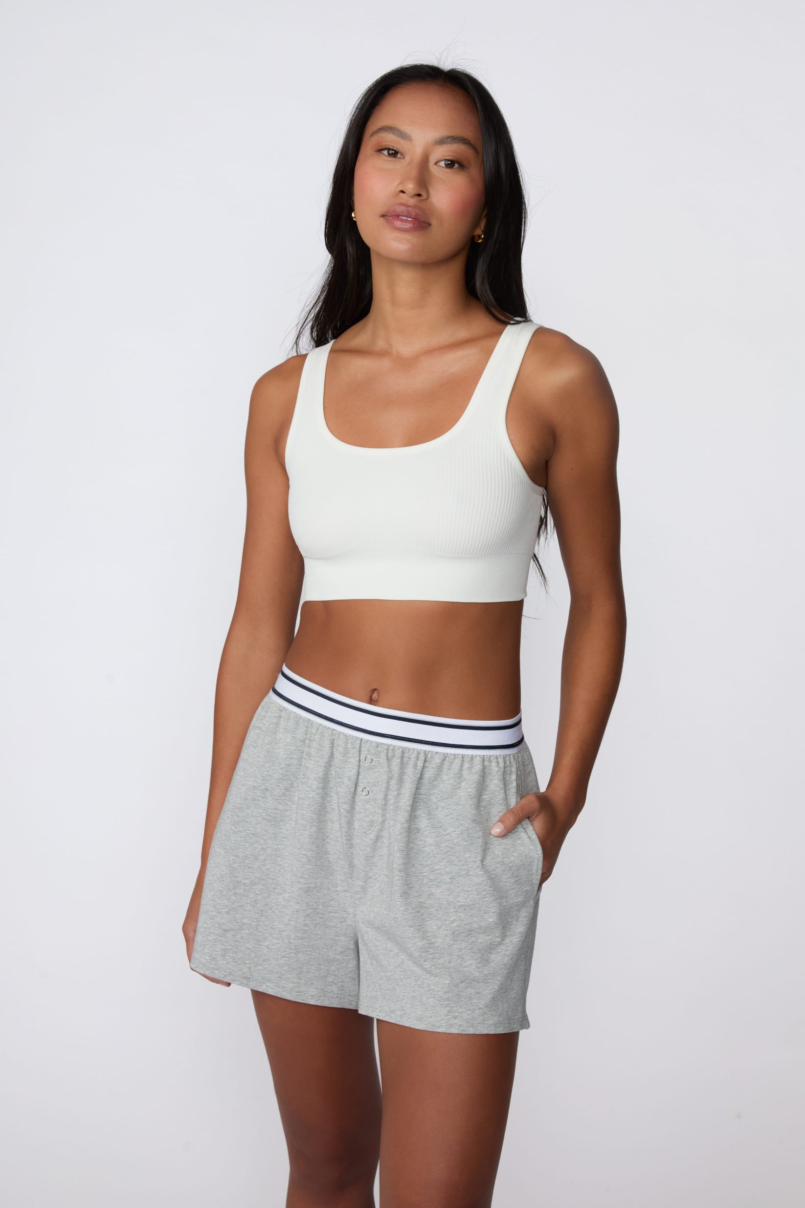 On a light gray studio backdrop, Amber wears the Cloud Cotton Pajama Short in Heather Grey with a white ribbed bralette. The full-body view showcases the relaxed fit, striped elastic waistband, side pockets, and easy sleepwear styling.