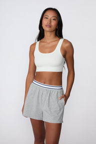 On a light gray studio backdrop, Amber wears the Cloud Cotton Pajama Short in Heather Grey with a white ribbed bralette. The full-body view showcases the relaxed fit, striped elastic waistband, side pockets, and easy sleepwear styling.