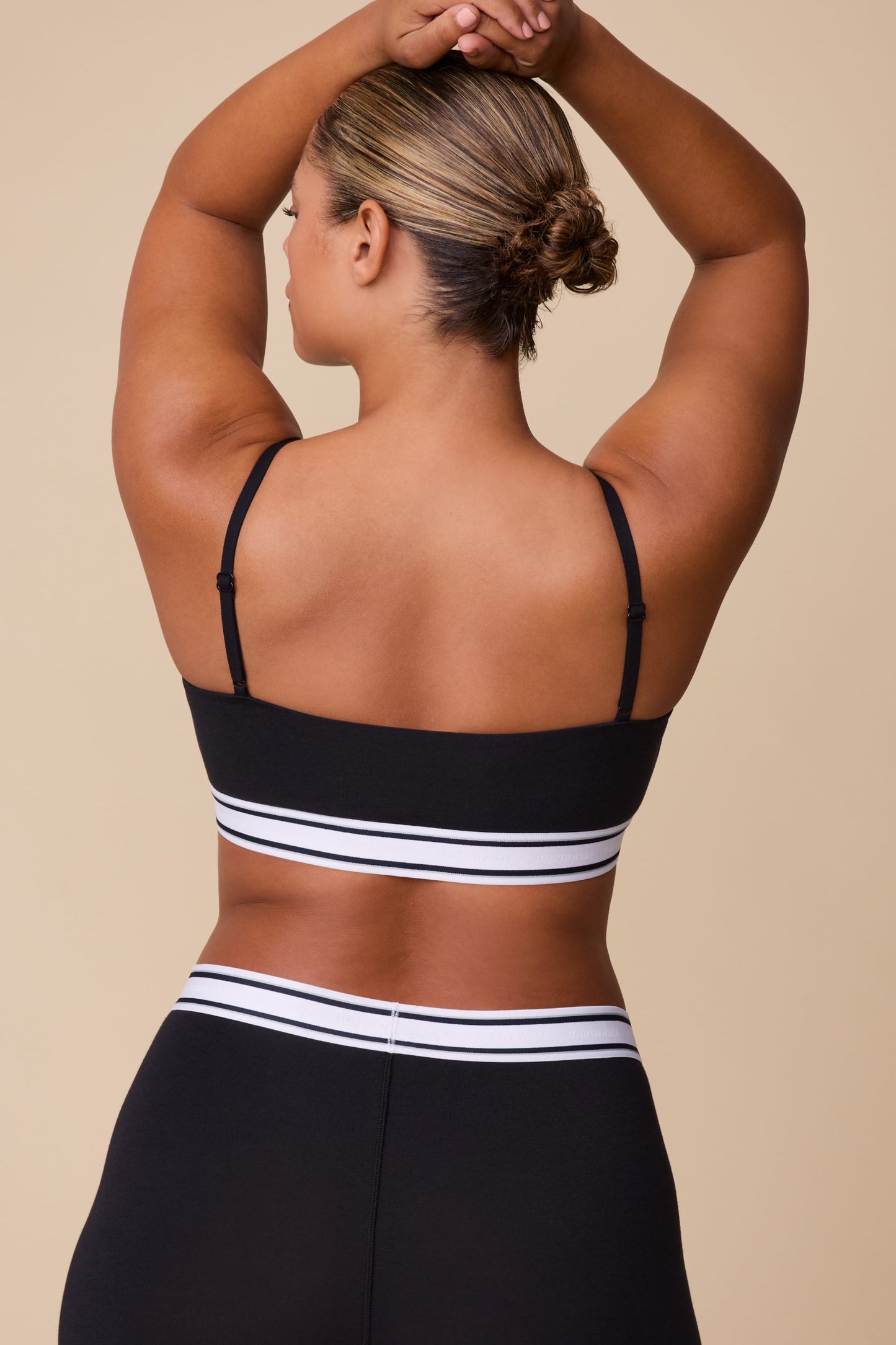 On a neutral beige backdrop, India wears the Cloud Cotton Retro Bandeau Bralette in Black with matching shorts, showcasing the back view, adjustable straps, and striped elastic underband.