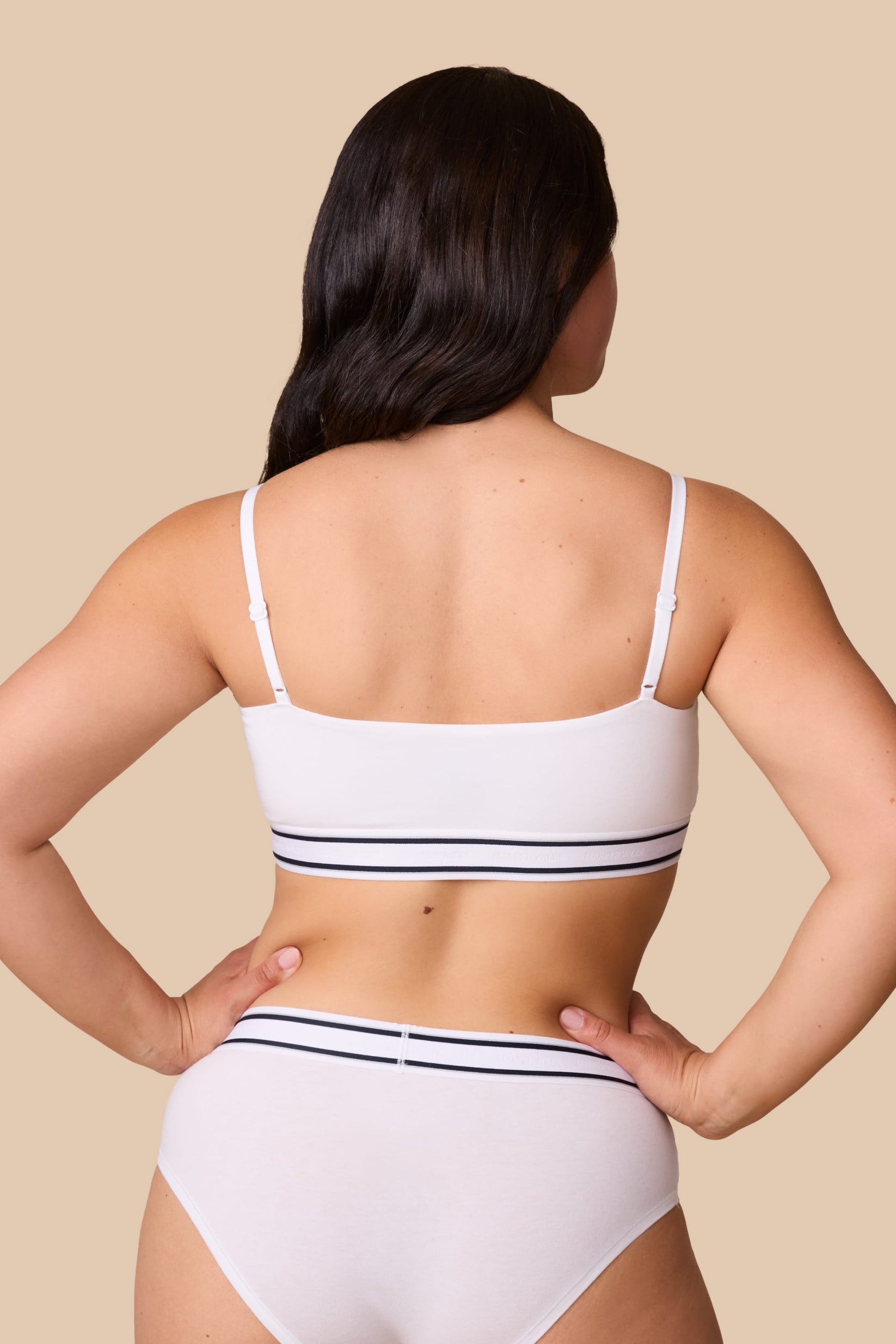 On a neutral beige backdrop, a back view of Emersyn wearing the Cloud Cotton Retro Bandeau Bralette in White with matching underwear highlights the straight back silhouette, adjustable straps, and striped elastic waistband details.