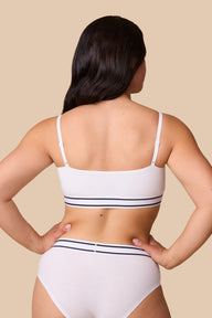 On a neutral beige backdrop, a back view of Emersyn wearing the Cloud Cotton Retro Bandeau Bralette in White with matching underwear highlights the straight back silhouette, adjustable straps, and striped elastic waistband details.