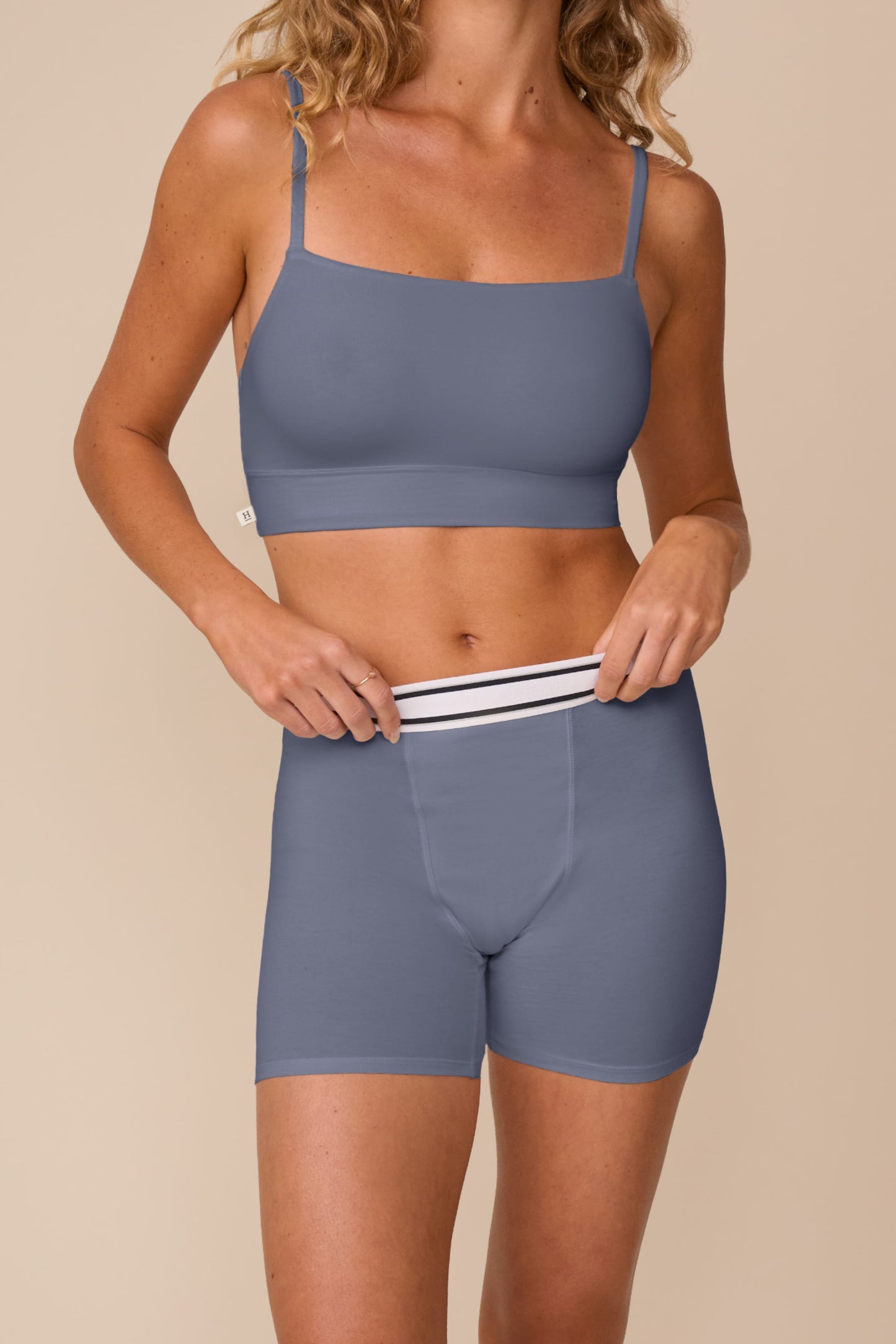 On a neutral studio backdrop, Freya wears the Cloud Cotton Retro Scoop Bralette and Cloud Cotton Retro Boxer Brief in Slate. She gently pulls the waistband of the boxer brief, highlighting the retro-inspired striped waistband, soft cotton modal fabric, and full-coverage boxer brief silhouette.