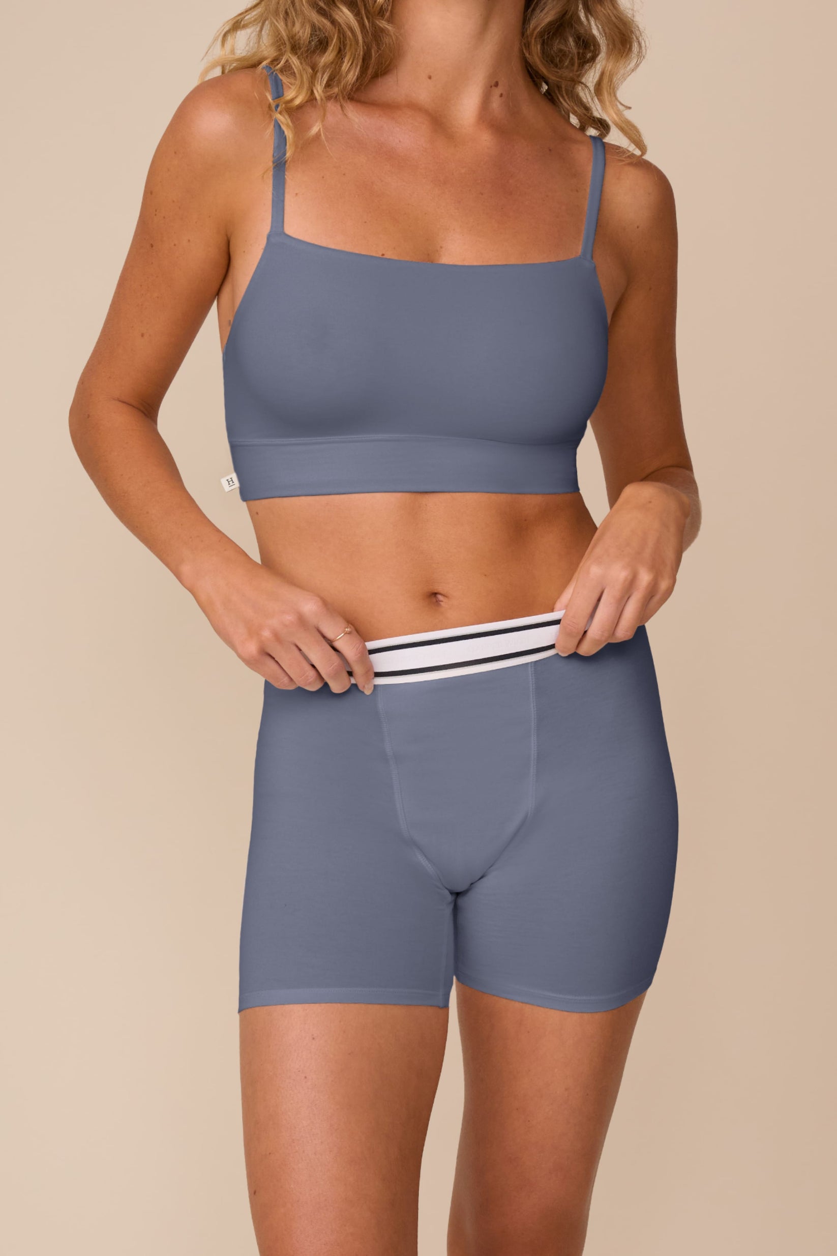 On a neutral studio backdrop, Freya wears the Cloud Cotton Retro Scoop Bralette and Cloud Cotton Retro Boxer Brief in Slate. She gently pulls the waistband of the boxer brief, highlighting the retro-inspired striped waistband, soft cotton modal fabric, and full-coverage boxer brief silhouette.