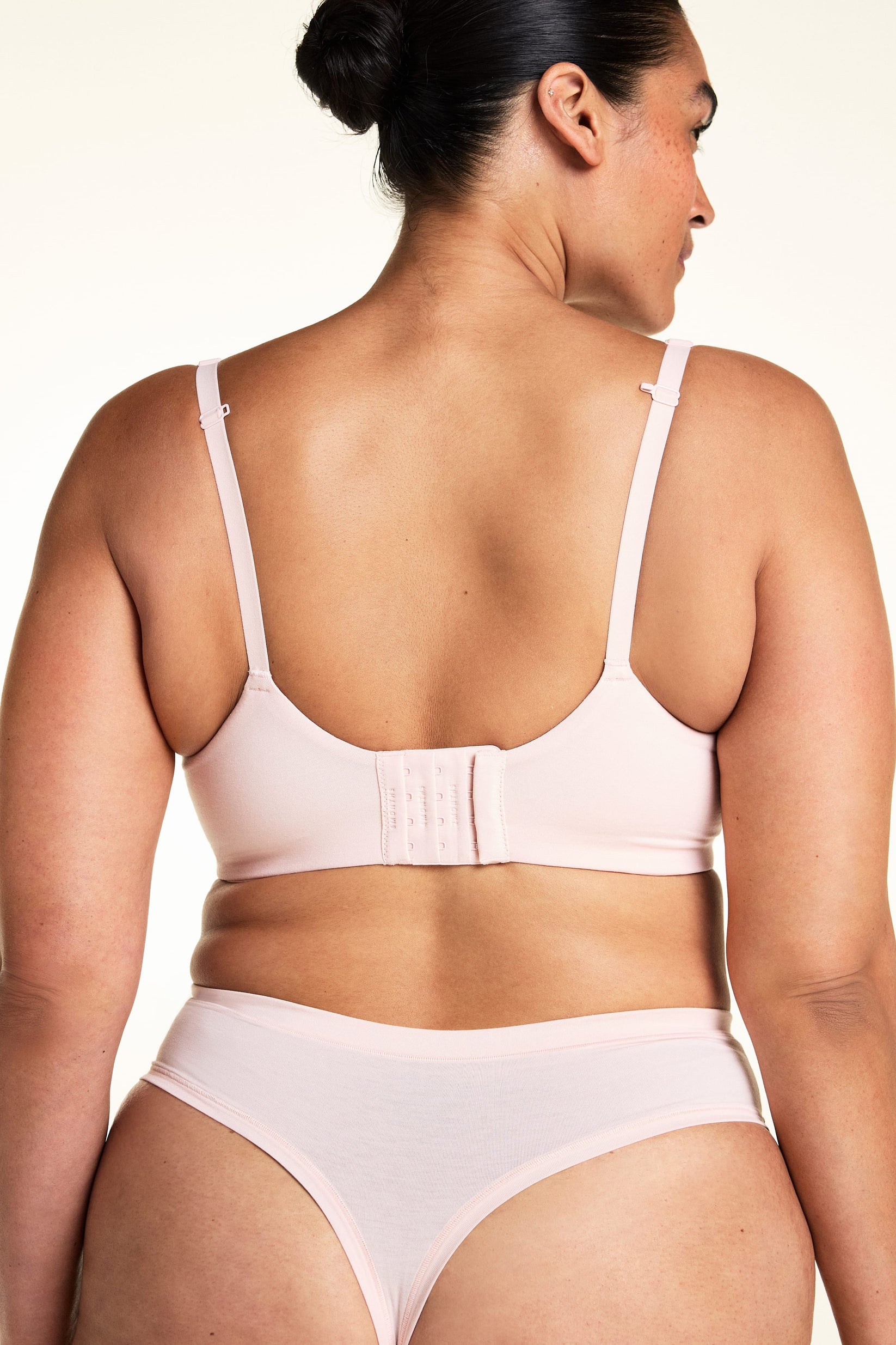 On a light studio backdrop, Jennifer wears the Cloud Cotton T-Shirt Bra in Dogwood paired with matching underwear. The angled back view showcases the convertible J-hook straps, adjustable fit, and smooth back closure.