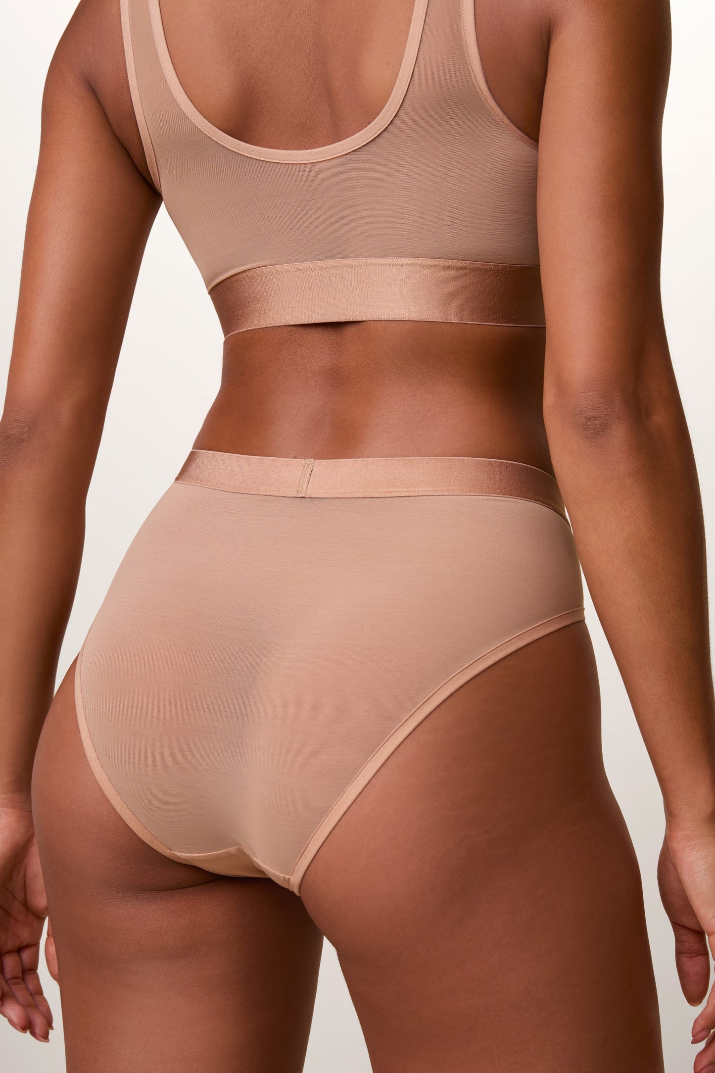 On a light studio backdrop, Ally wears the ComfortMesh™ Scoop Bralette and ComfortMesh™ Highwaist Brief in Taupe. The back view highlights the sheer mesh construction, moderate back coverage, and smooth waistband.