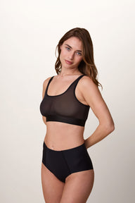 On a light studio backdrop, Gracie wears the ComfortMesh™ Scoop Bralette in Black with the matching high-waisted brief. A side-angle view showcases the scoop neckline, sheer mesh fabric, and smooth silhouette.
