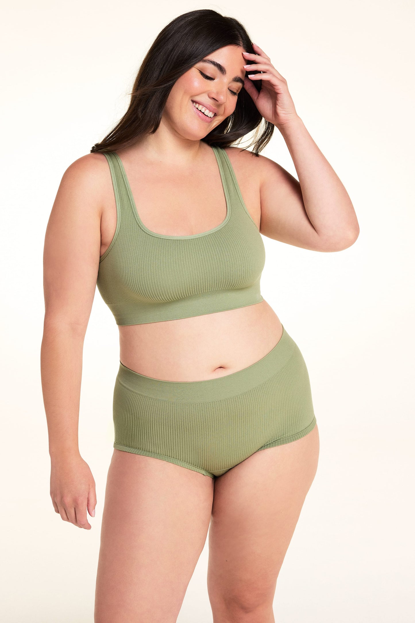 On a white studio backdrop, Alessandra wears the Lounge Rib Scoop Bralette in Dill with the matching Lounge Rib Sleepshort, smiling with one hand in her hair. The angled front view highlights the scoop neckline, ribbed fabric, and wide underband.