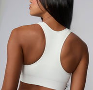 On a neutral studio backdrop, Amber is shown from the back wearing the Seamless Racerback Sports Bra in White. The racerback silhouette, breathable textured panels, and wide wireless band are clearly visible.