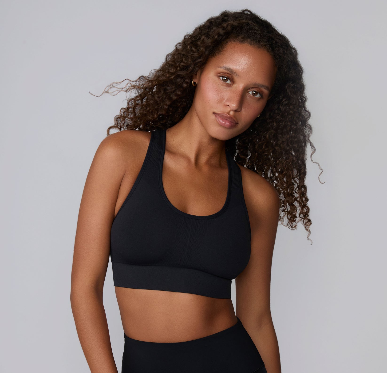 On a neutral studio backdrop, Courtney is shown in a front-facing close-up wearing the Seamless Racerback Sports Bra in Black. The smooth scoop neckline, wireless support, and wide underband are visible as she tilts her head slightly.