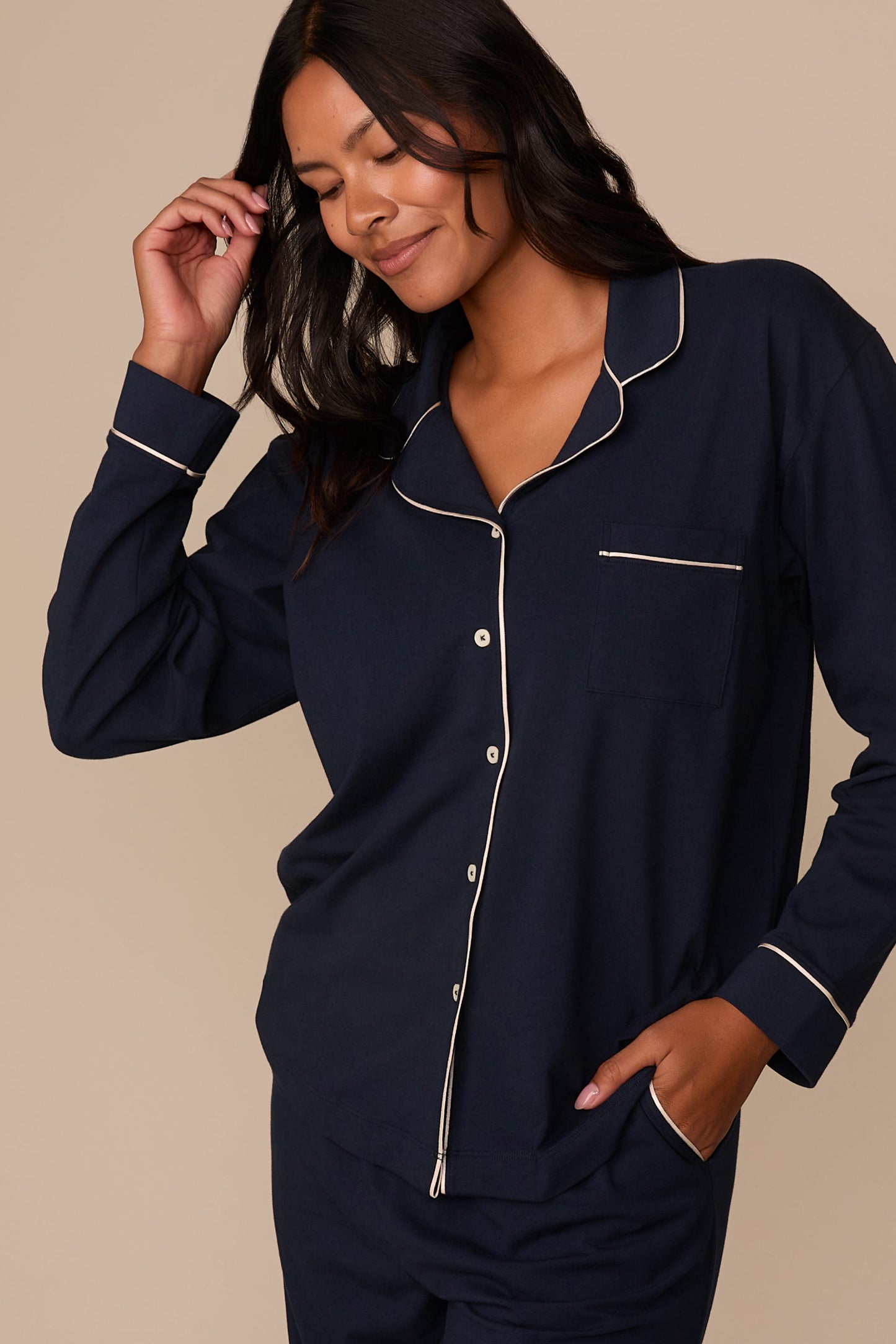 On a neutral studio backdrop, Victoria wears the Perfect Pima Pajama Long Sleeve Shirt in Navy with matching pajama pants. The angled view showcases the relaxed fit, chest pocket, button-front closure, side pocket detail on the pants, and contrast piping along the collar, placket, pocket, and cuffs.