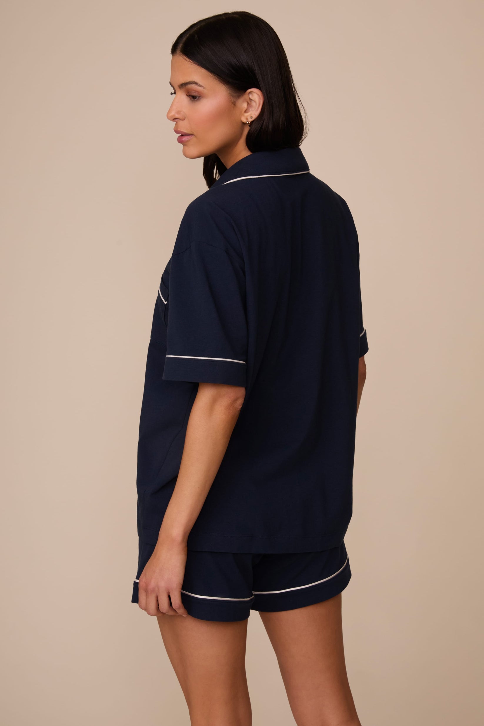 On a neutral studio backdrop, Marcela wears the Perfect Pima Pajama Shirt in Navy with matching navy pajama shorts. The back view showcases the relaxed fit, short sleeves, and contrast piping along the collar and sleeve cuffs.