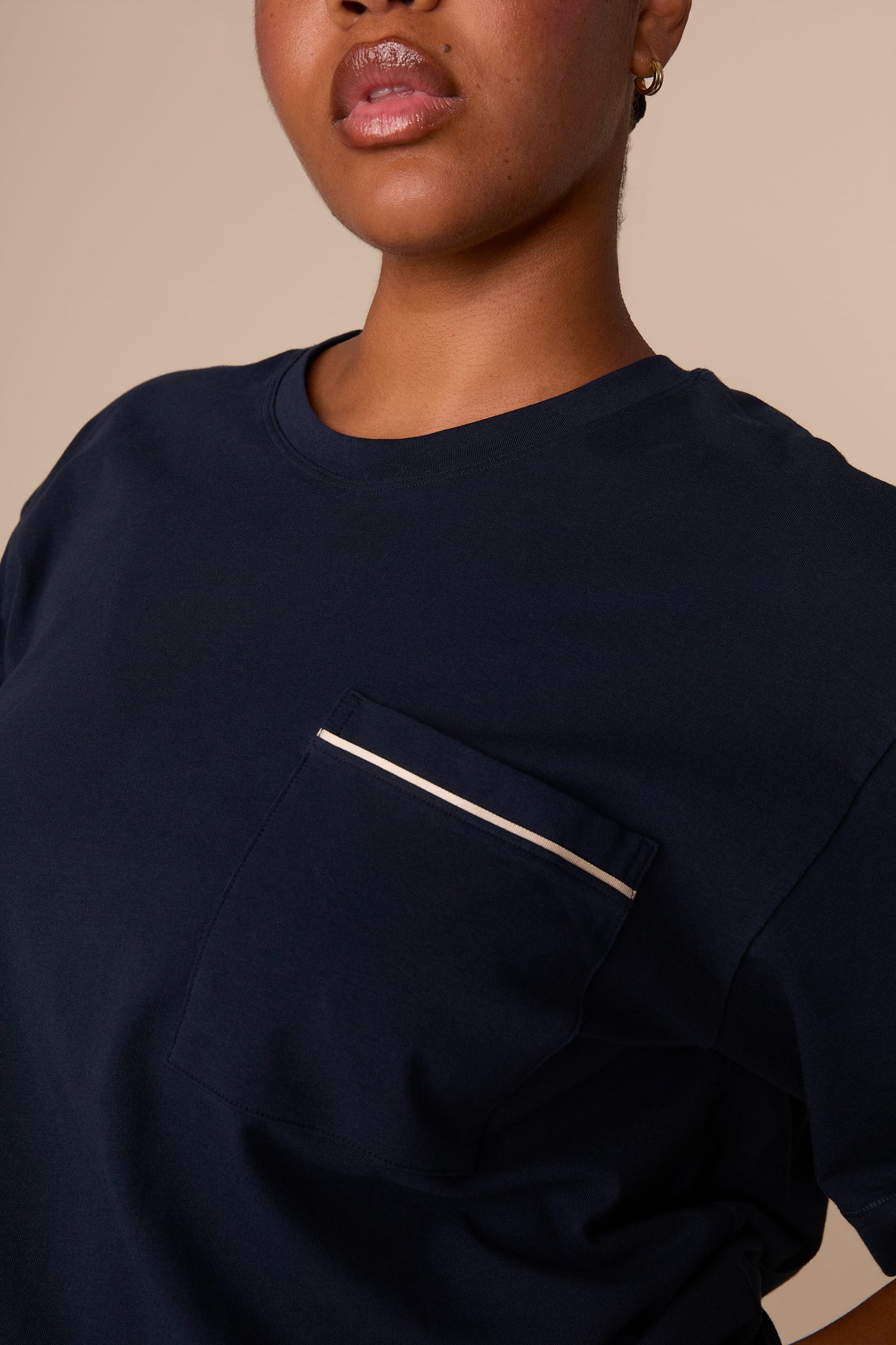 On a warm beige studio backdrop, Zarria wears the Perfect Pima Pajama T-Shirt in a deep navy color. The close-up detail view highlights the crew neckline and chest pocket finished with contrasting piping.