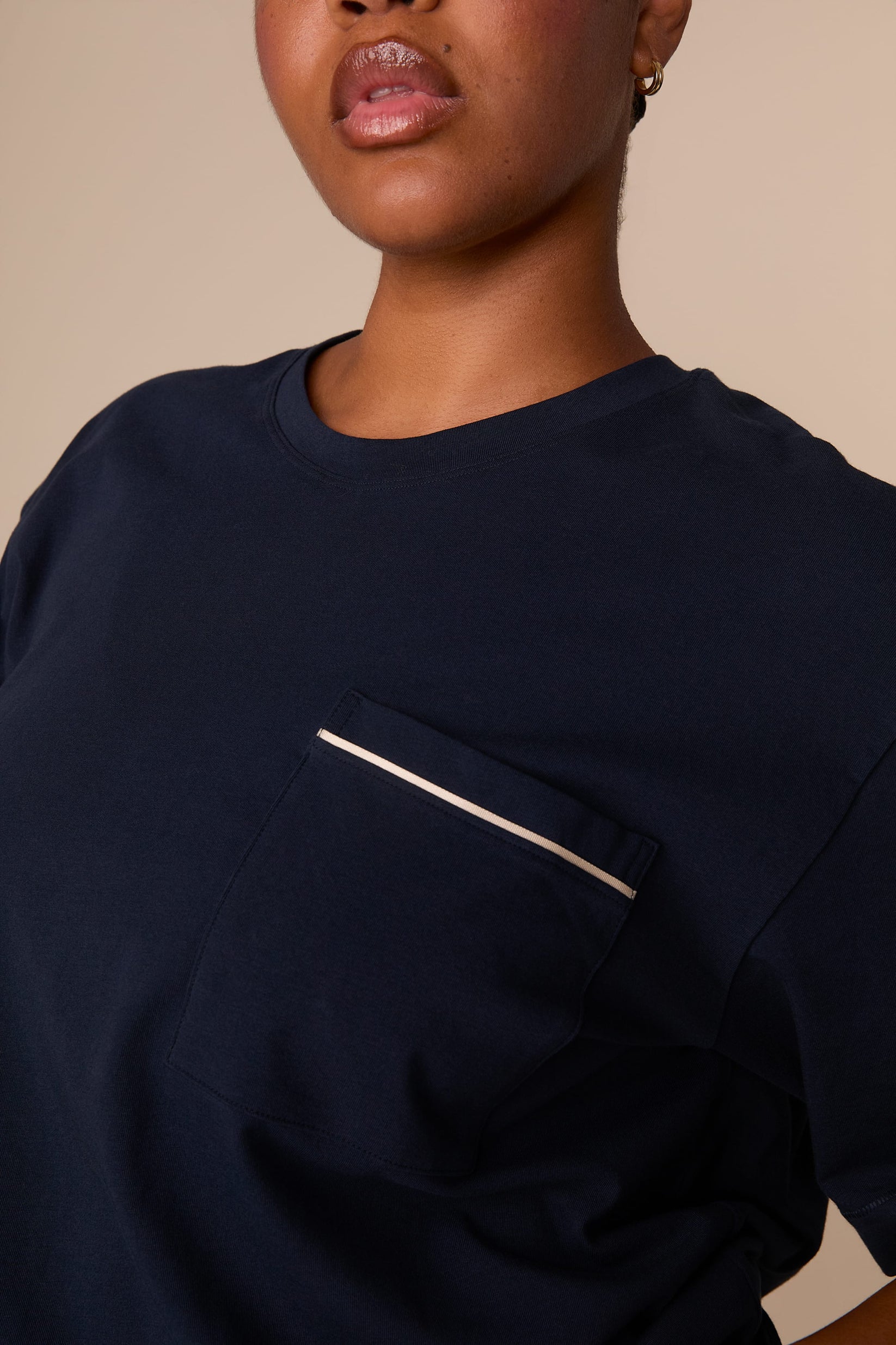On a warm beige studio backdrop, Zarria wears the Perfect Pima Pajama T-Shirt in a deep navy color. The close-up detail view highlights the crew neckline and chest pocket finished with contrasting piping.