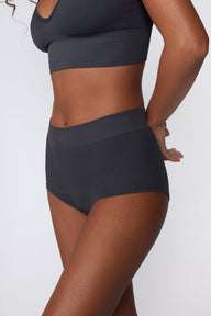 In a studio setting, Courtney wears the Ribbed Seamless Boyshort in Charcoal with a matching ribbed bralette. A side-angle close-up highlights the high-rise waistband, boyshort silhouette, and ribbed texture.