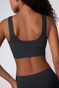 On a light gray studio backdrop, Courtney wears the Ribbed Seamless Scoop Bralette in Charcoal. A back view highlights the scoop-back design, wide straps, ribbed texture, and supportive underband.