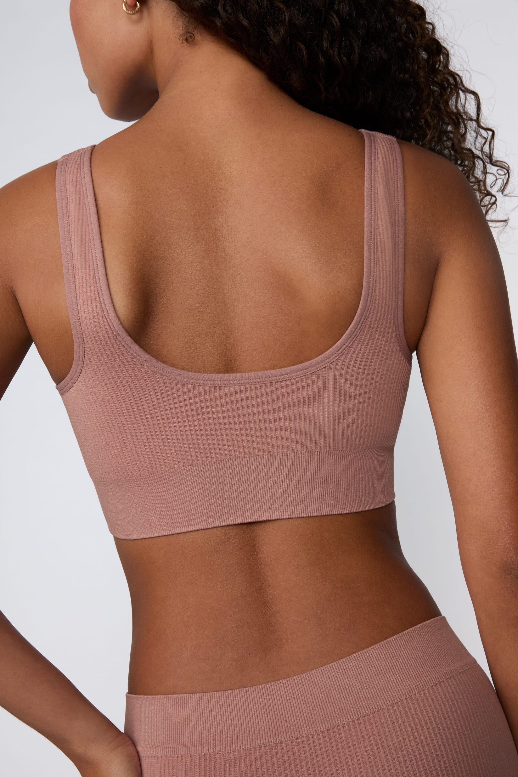 On a light gray studio backdrop, Courtney wears the Ribbed Seamless Scoop Bralette in Plum. A back view highlights the scoop-back design, wide straps, ribbed texture, and supportive underband.