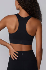 On a light gray studio backdrop, Courtney wears the Seamless Racerback Sports Bra in Black with matching bike shorts. The back view showcases the racerback design, breathable knit texture, wide underband, and tonal Harper Wilde logo at the center back.